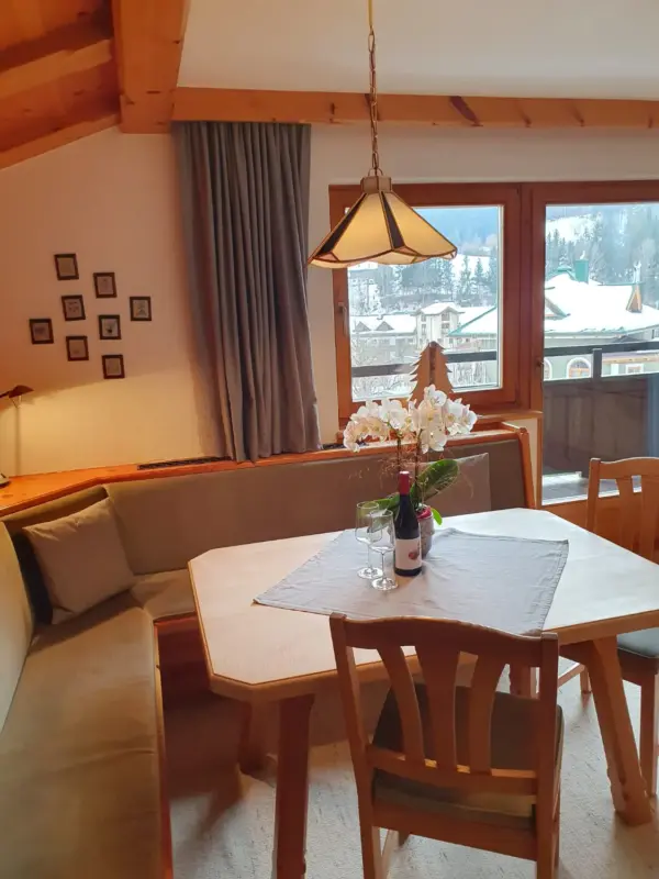 Gemütliche Essecke im rustikalen Tiroler Stil mit gepolsterter Eckbank und Holztisch. Auf dem Tisch stehen Weingläser und Orchideen, im Hintergrund der Blick durch das Fenster auf das verschneite Fieberbrunn.